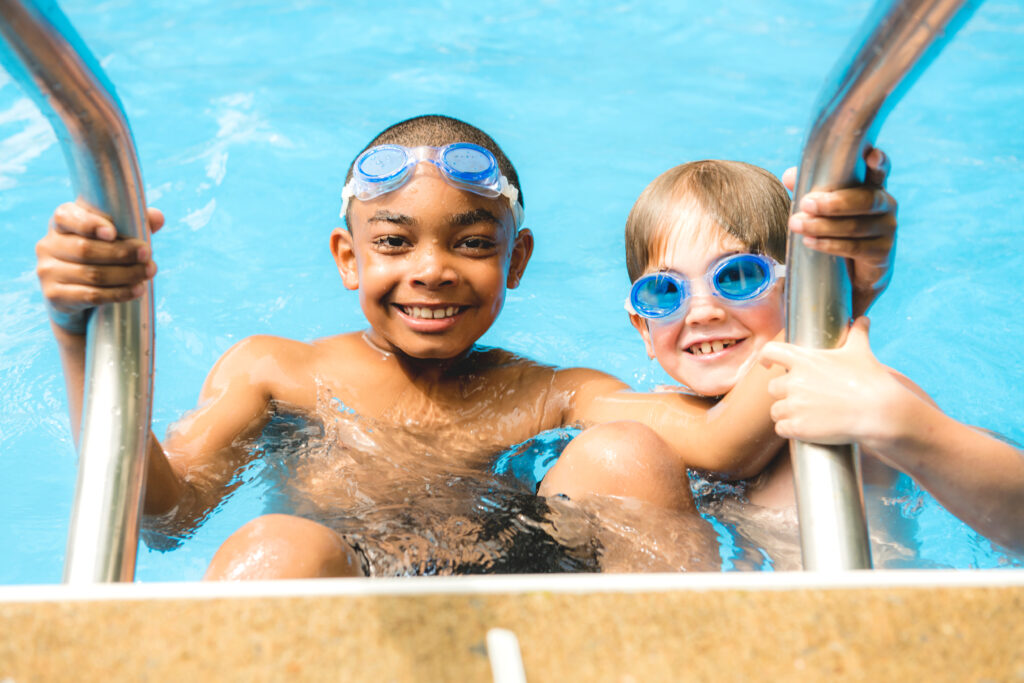 Two little kids holding onto the railing of the pool, smiling.