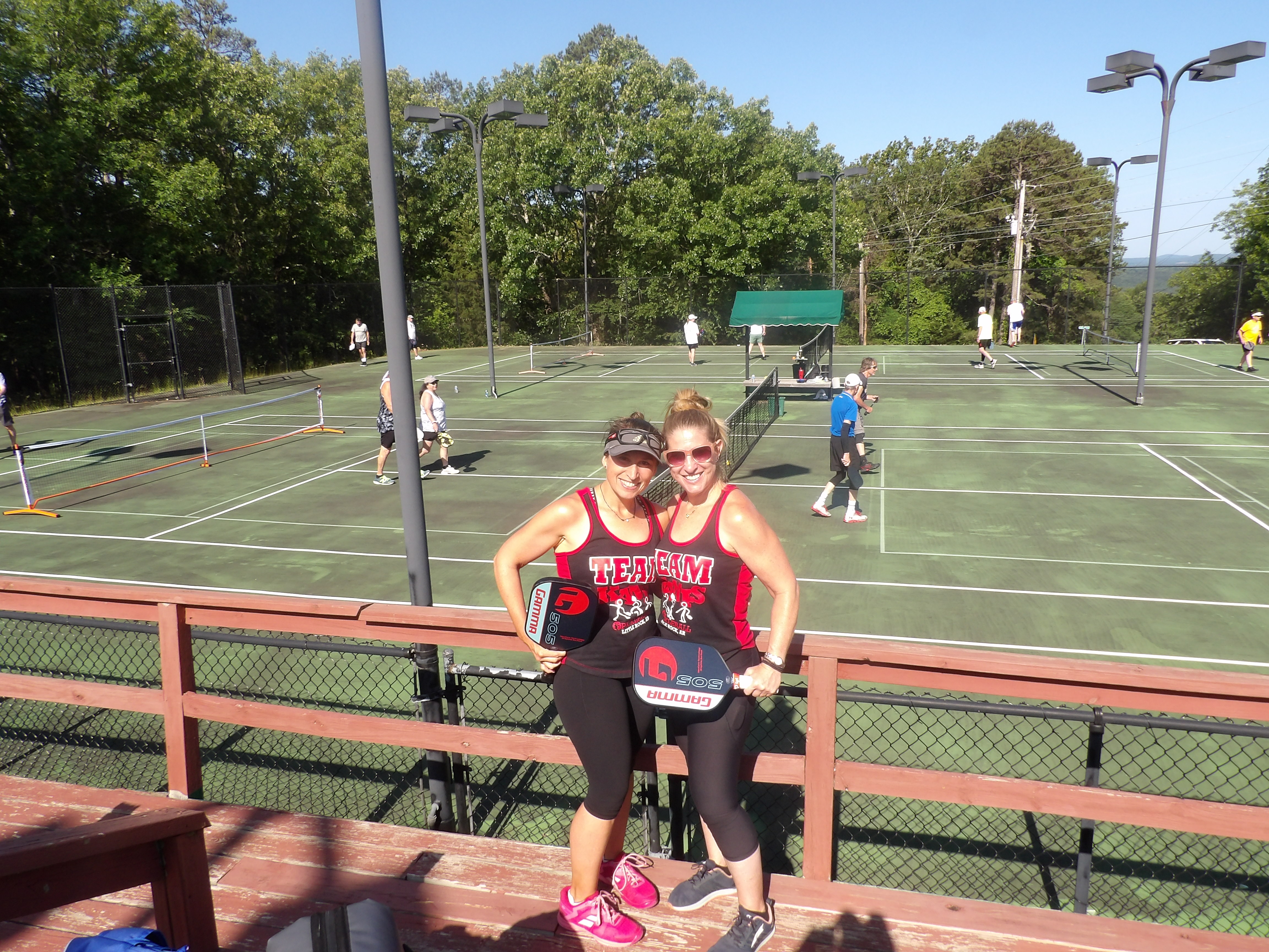 Two women in matching tank tops and Pickleball paddles posed in front of Pickleball courts in Fairfield Bay, AR.